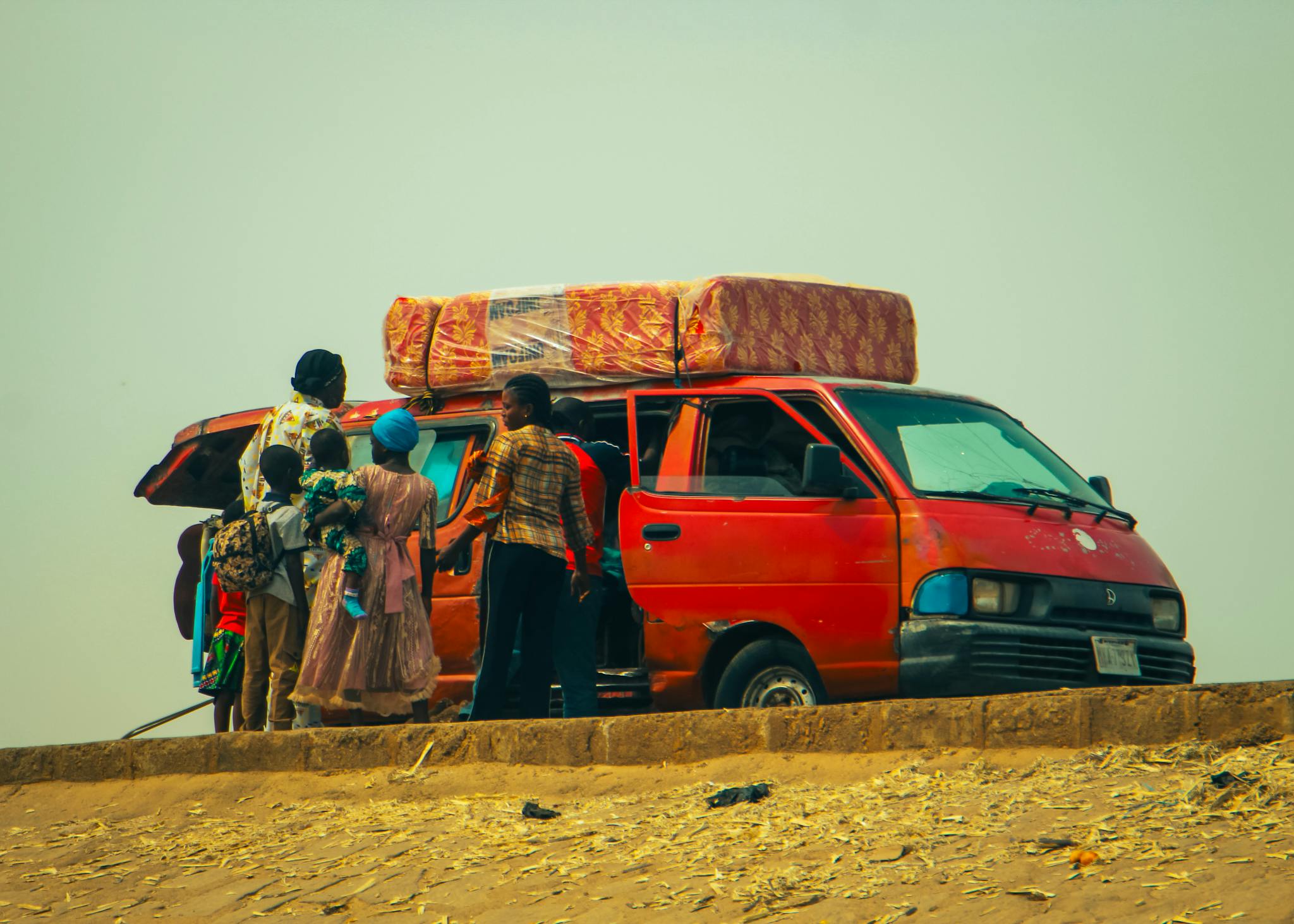 A family gathers beside a red van loaded with luggage on a sandy road, showcasing travel and adventure.
